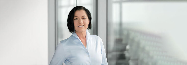 Eine Frau mit dunklen Haaren und heller Bluse steht vor einem Fenster im Büro.